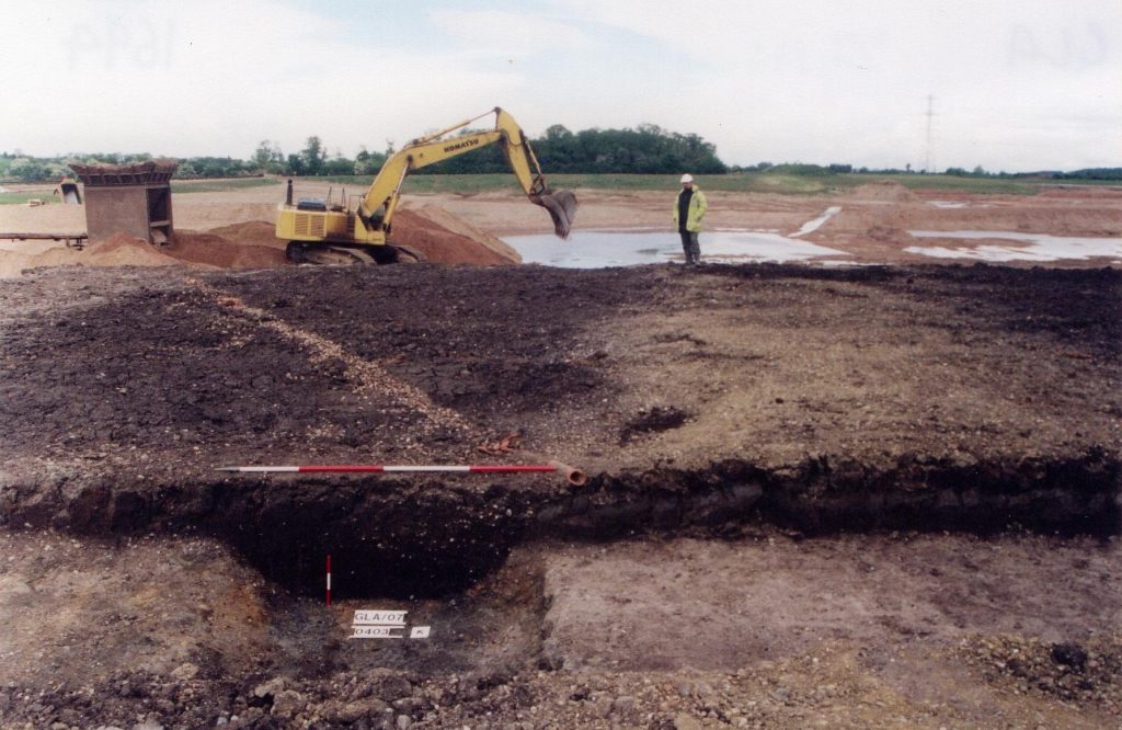 A photo of a large archaeological site. A man in a hard hat stands next to a yellow digger.