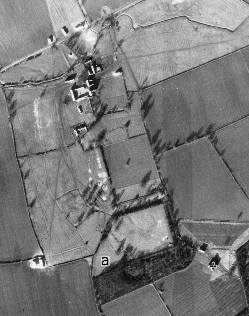A black and white aerial photo of a complex of features, including buildings, earthworks and fields.