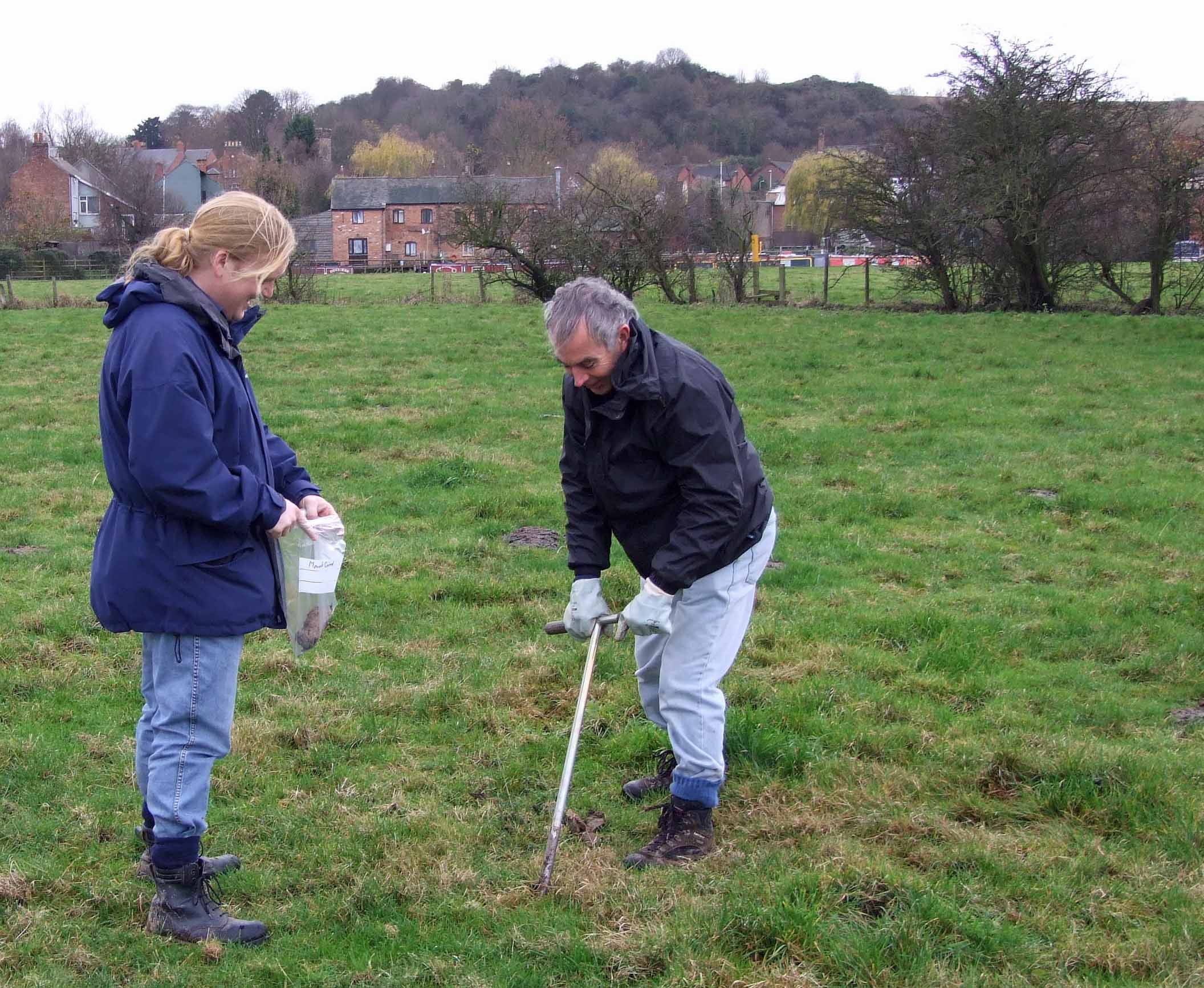 A photograph of two people working in a field. Hills, trees and buildings can be seen in the distance. One person uses a tool to collect a sample from the ground while another holds a bag open for the sample to be collected.