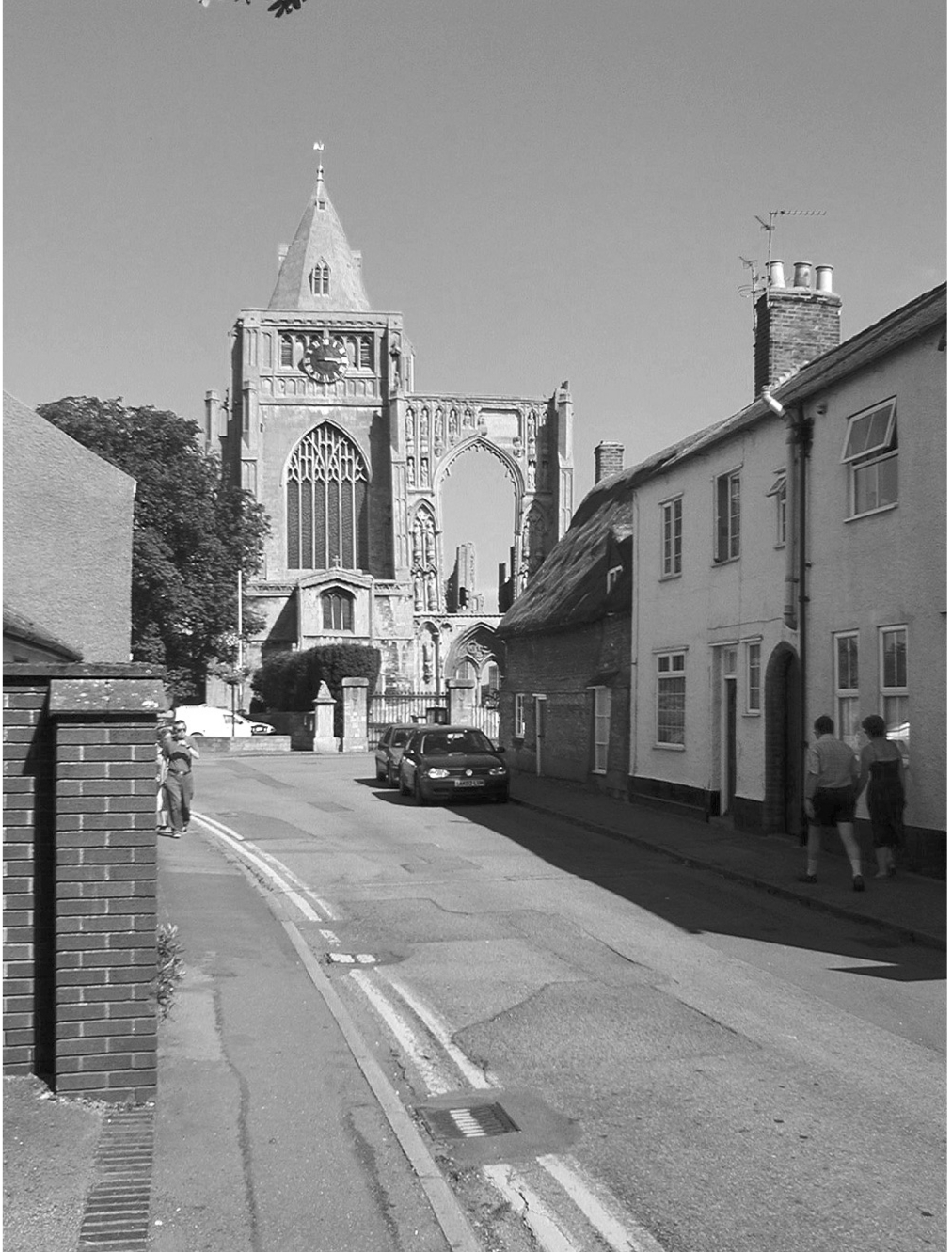 A black and white photograph of a street. There are houses on the right hand side, and at the end is a partially ruined medieval abbey.