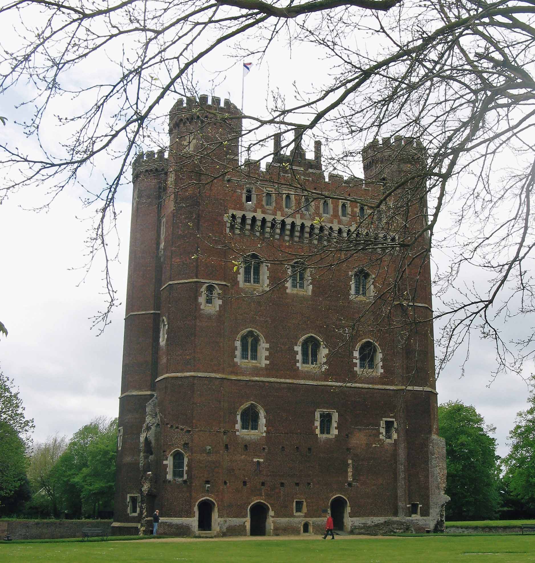 A photo of a tall, turreted castle, surrounded by grass and trees. It built out of red bricks, with pale stone details round the windows and doors.  