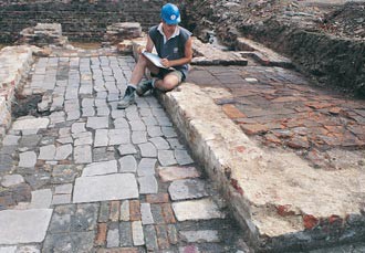 Archaeologist sat on a wall with a clipboard, looking at an exposed floor.