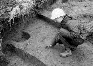 An archaeologist with a towel in their hand, crouching in a trench with a semi circular feature in front of them.