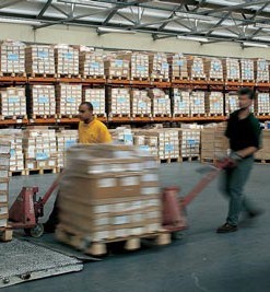 A warehouse, with two people moving boxes on trolleys.